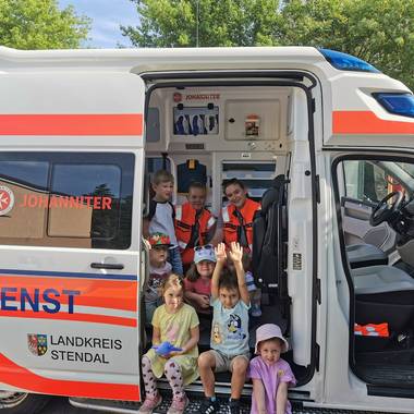 Ein Rettungswagen steht mit geöffneter Seitentür, zusehen sind zwei Rettungssanitäterinnen und davor mehrere Kinder.